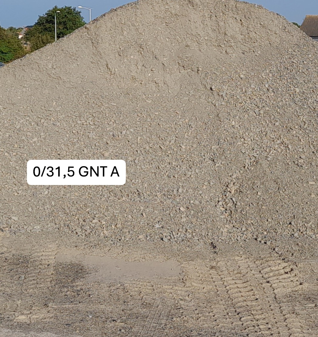 Tas de gravier gris étiqueté "0/31,5 GNT A" au centre, posé sur un sol compacté avec des traces de machines visibles en bas; arbres en arrière-plan à gauche.