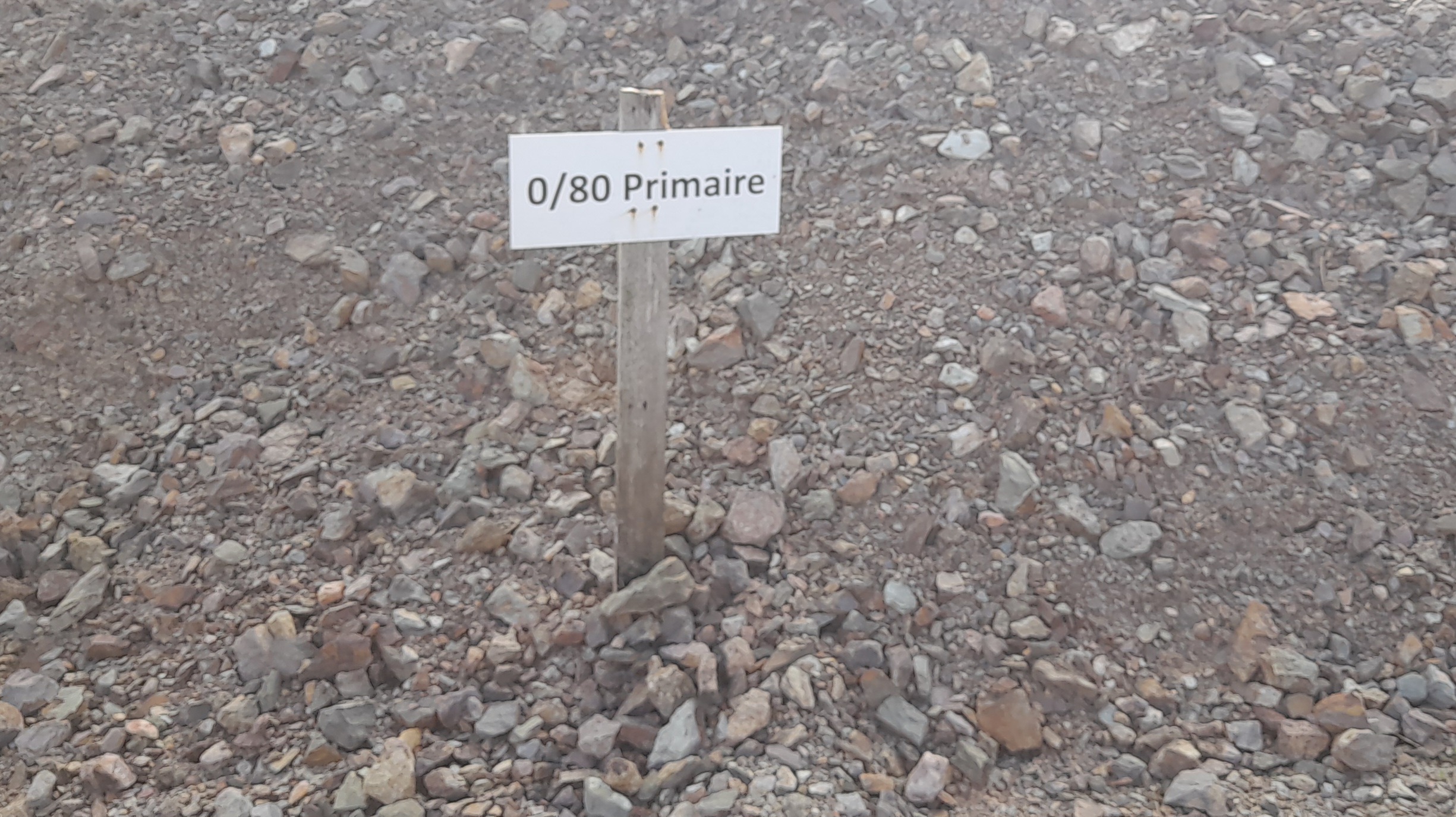 Panneau en bois planté dans un tas de gravier, avec une inscription "0/80 Primaire" indiquant la taille et le type de granulats. Le gravier composé de pierres de tailles variées entoure le panneau sur toute l'image.