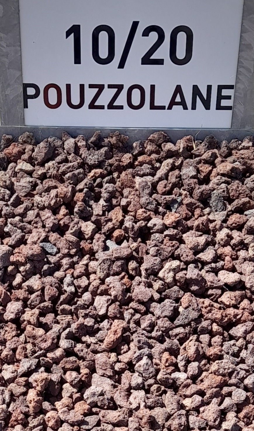 Gravier rouge-brun de pouzzolane avec un panneau indiquant "10/20 POZZOLANE" en arrière-plan, représentant des granulats de taille 10 à 20 mm.