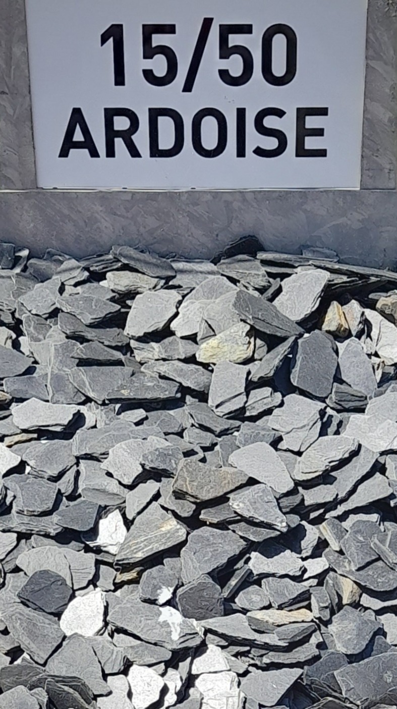 Tas d'éclats d'ardoise gris foncé avec une pancarte au sommet indiquant "15/50 ARDOISE", représentant la taille des morceaux d'ardoise présentés pour des travaux de décoration ou de construction.