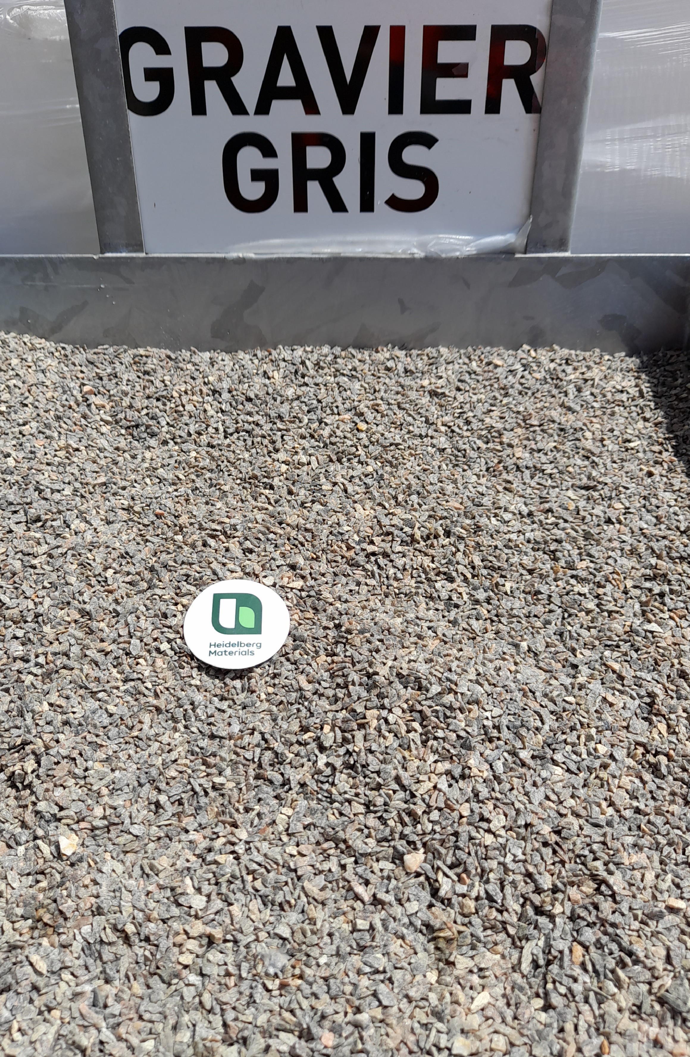 Tas de gravier gris avec un panneau blanc en arrière-plan indiquant "GRAVIER GRIS" en lettres noires. Un disque blanc marqué d'un logo vert est placé au centre du tas.