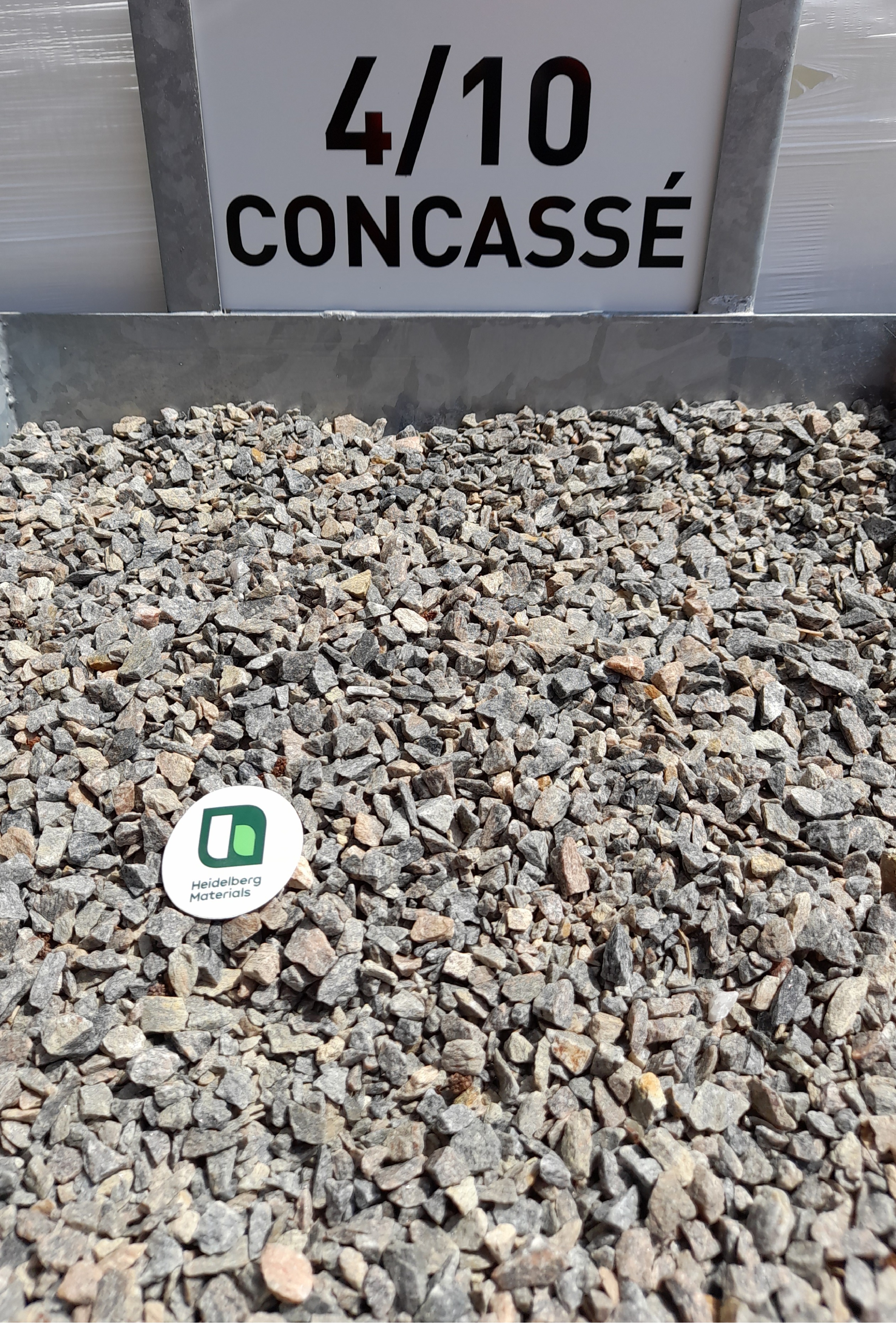 Gravier concassé de taille 4/10 visible en gros plan, avec un échantillon exposé dans un bac métallique. Une étiquette verte et blanche est posée au centre pour identifier le produit.