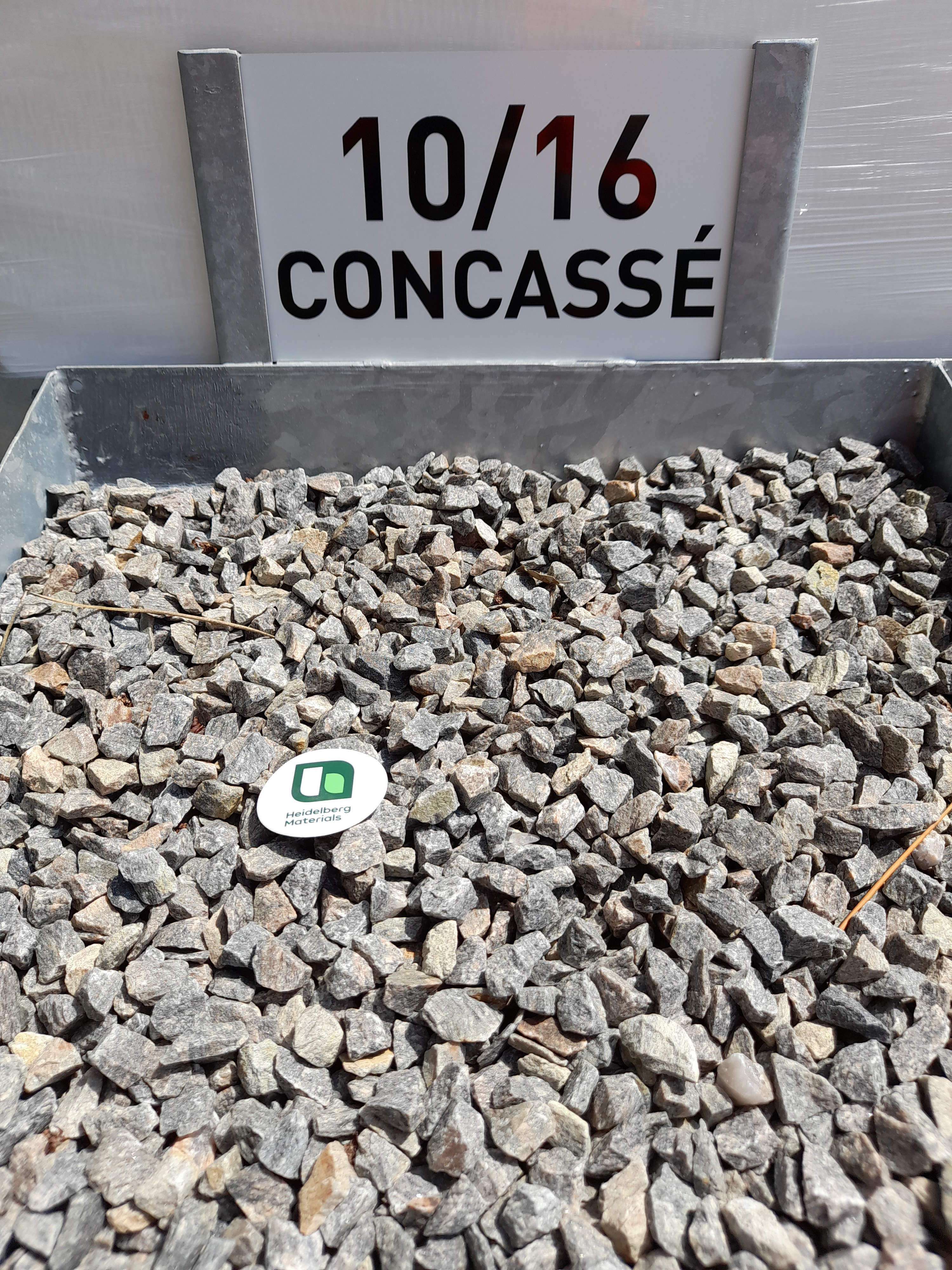 Gravier concassé gris de calibre 10/16 présenté dans un bac métallique, avec une étiquette circulaire blanche portant un logo vert placé au centre. Panneau à l'arrière indiquant "10/16 CONCASSÉ".