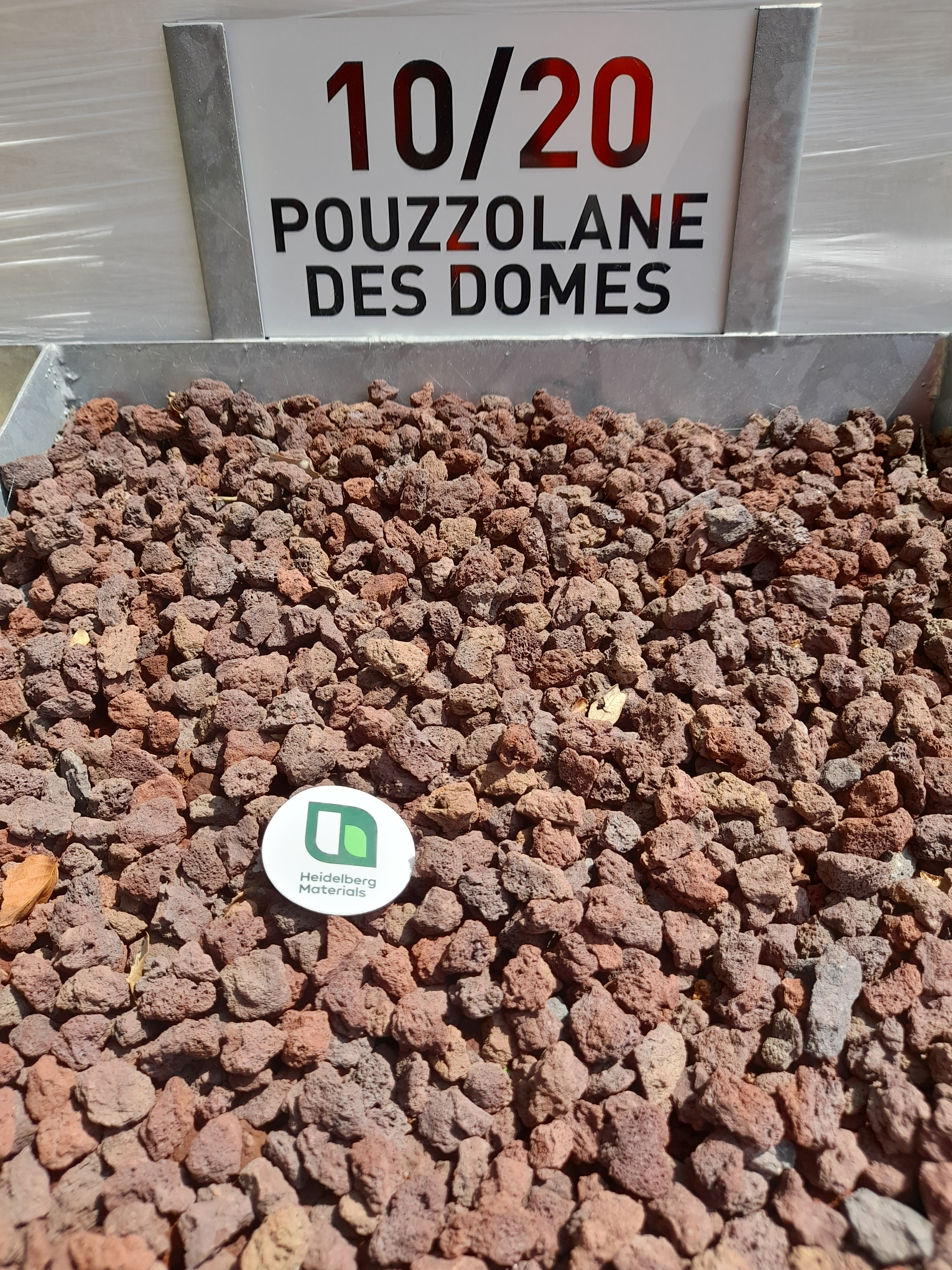 Graviers rouges de pouzzolane disposés dans un bac métallique, avec un panneau en arrière-plan indiquant "10/20 Pouzzolane des Dômes". Un petit disque blanc avec un logo vert est visible au centre des graviers.