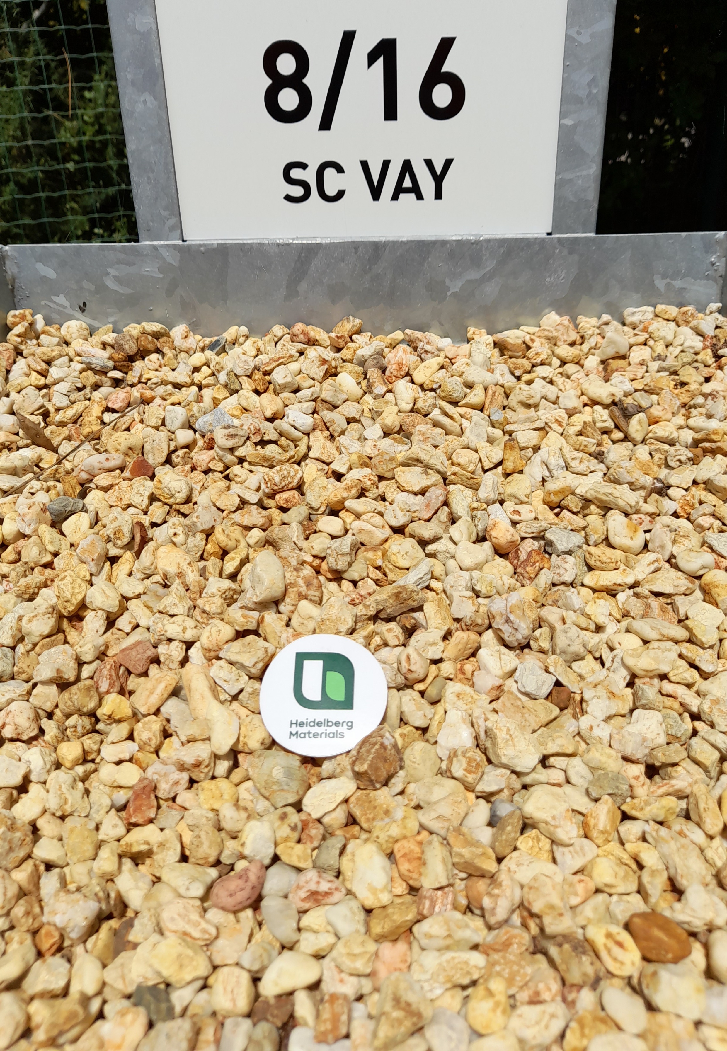 Tas de graviers de couleur beige doré avec une étiquette ronde blanche au centre affichant un logo vert et blanc. En arrière-plan, un panneau gris indique "8/16 SC VAY".