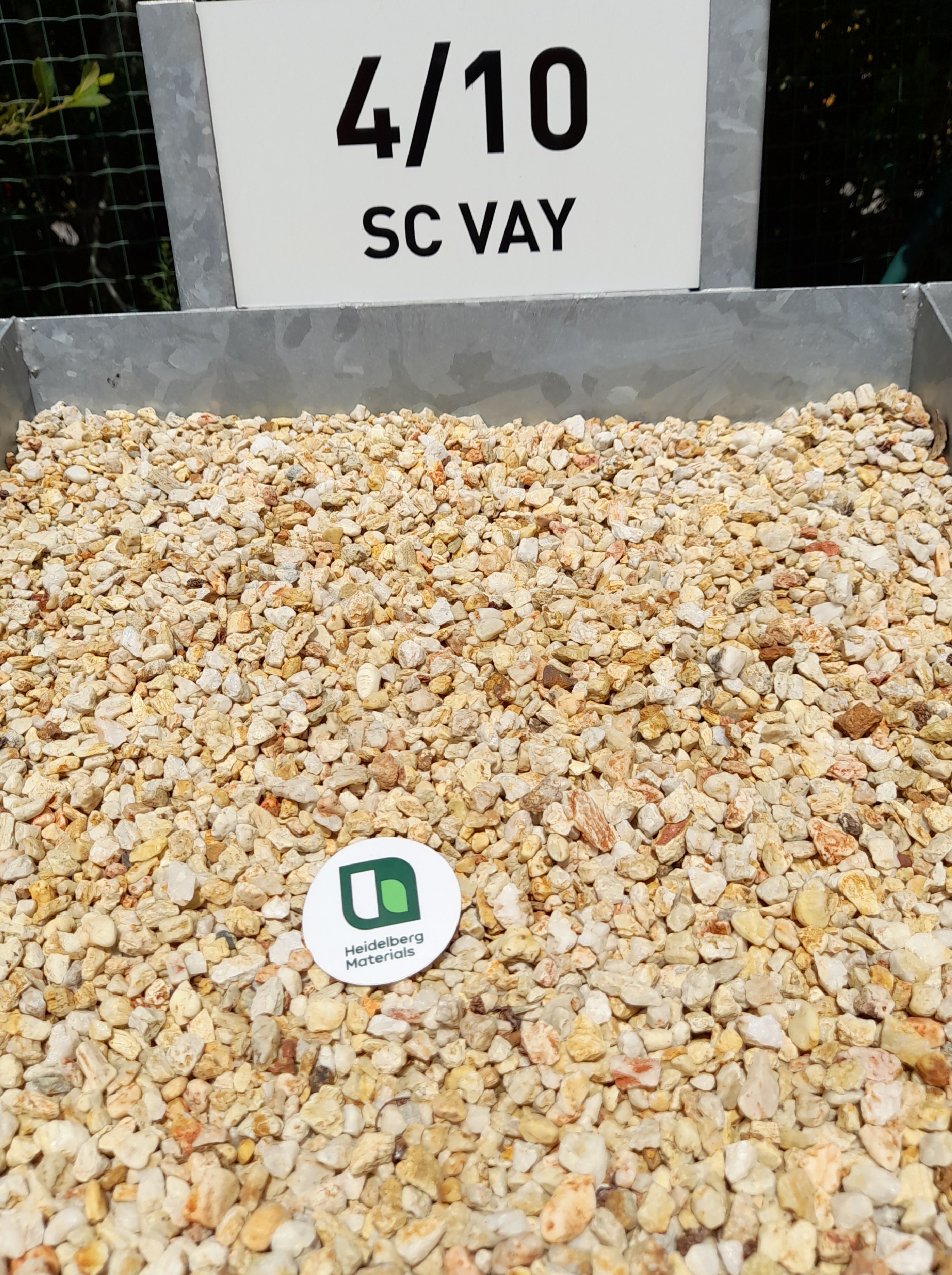 Tas de gravillons de granulométrie 4/10, identifié par un panneau blanc avec l'inscription "4/10 SC VAY" en haut, et un disque publicitaire vert et blanc au centre du gravier.