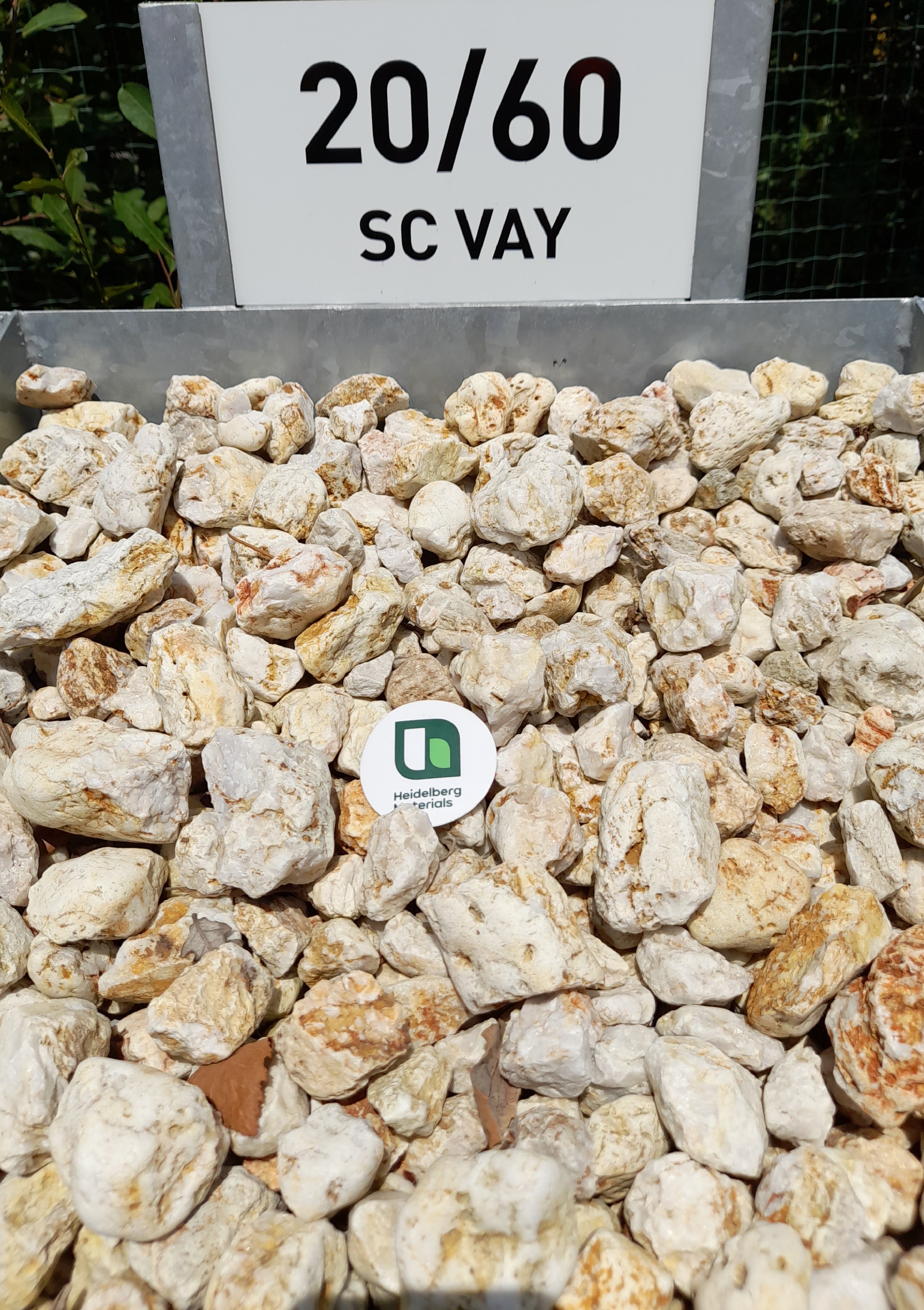Tas de graviers blancs et beige de taille 20/60 mm, marqué par une pancarte blanche indiquant "20/60 SC VAY" en haut de l'image. Un jeton vert avec un logo est posé au centre des graviers. Fond verdoyant en arrière-plan.