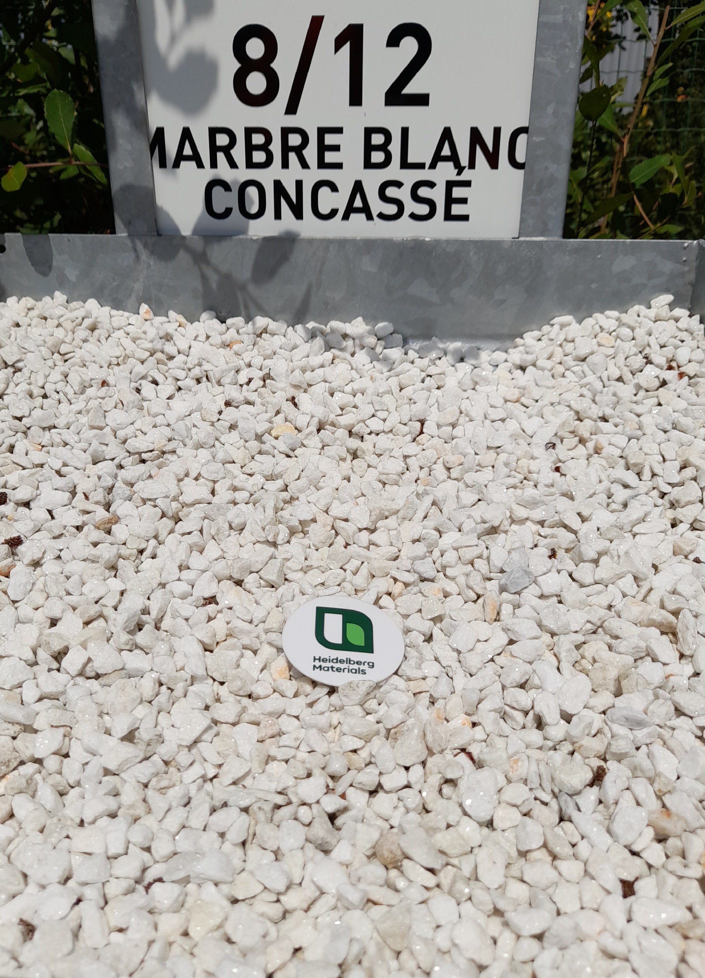 Tas de gravier de marbre blanc concassé, granulat de taille 8/12 mm indiqué par un panneau en arrière-plan. Un petit badge rond avec un logo vert et noir est posé au centre du gravier.