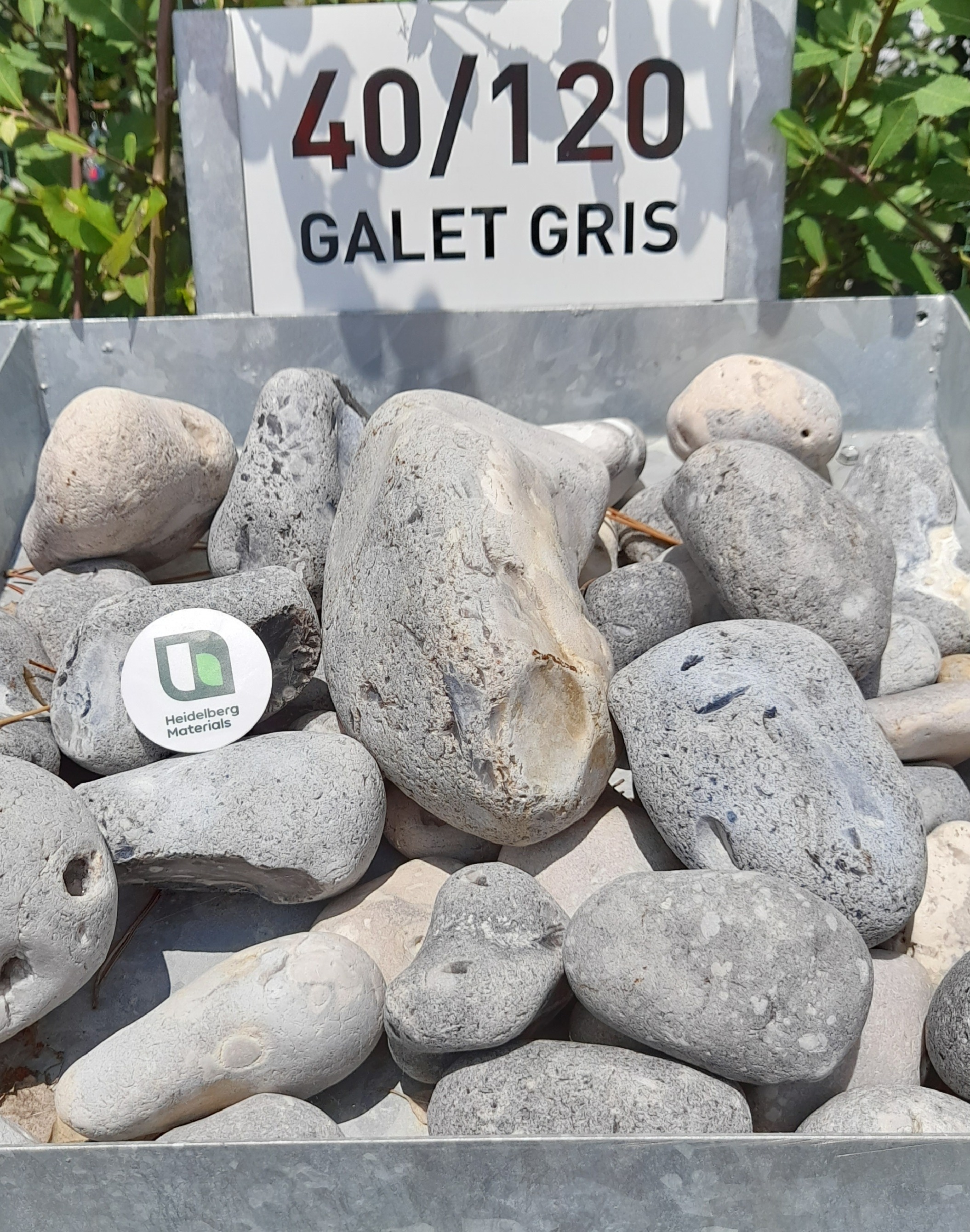 Galets gris de différentes tailles (40/120 mm) disposés dans un bac métallique, étiquetés avec un panneau. Fond de feuillage vert visible en arrière-plan.