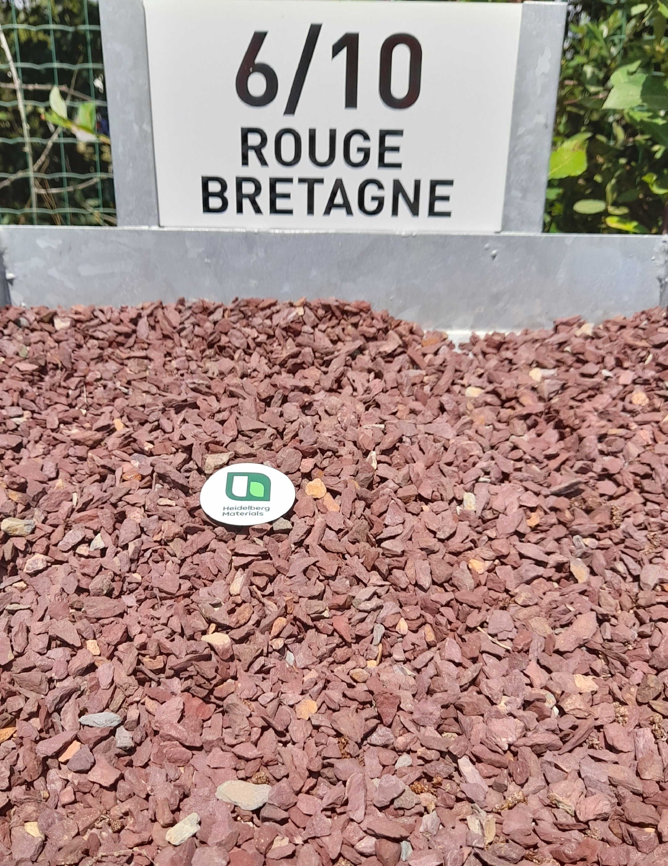Petit gravier rouge étiqueté “6/10 ROUGE BRETAGNE” en arrière-plan, avec un disque blanc portant un code QR posé au centre de la surface des gravillons rouges. Fond végétal flou visible en haut de l’image.