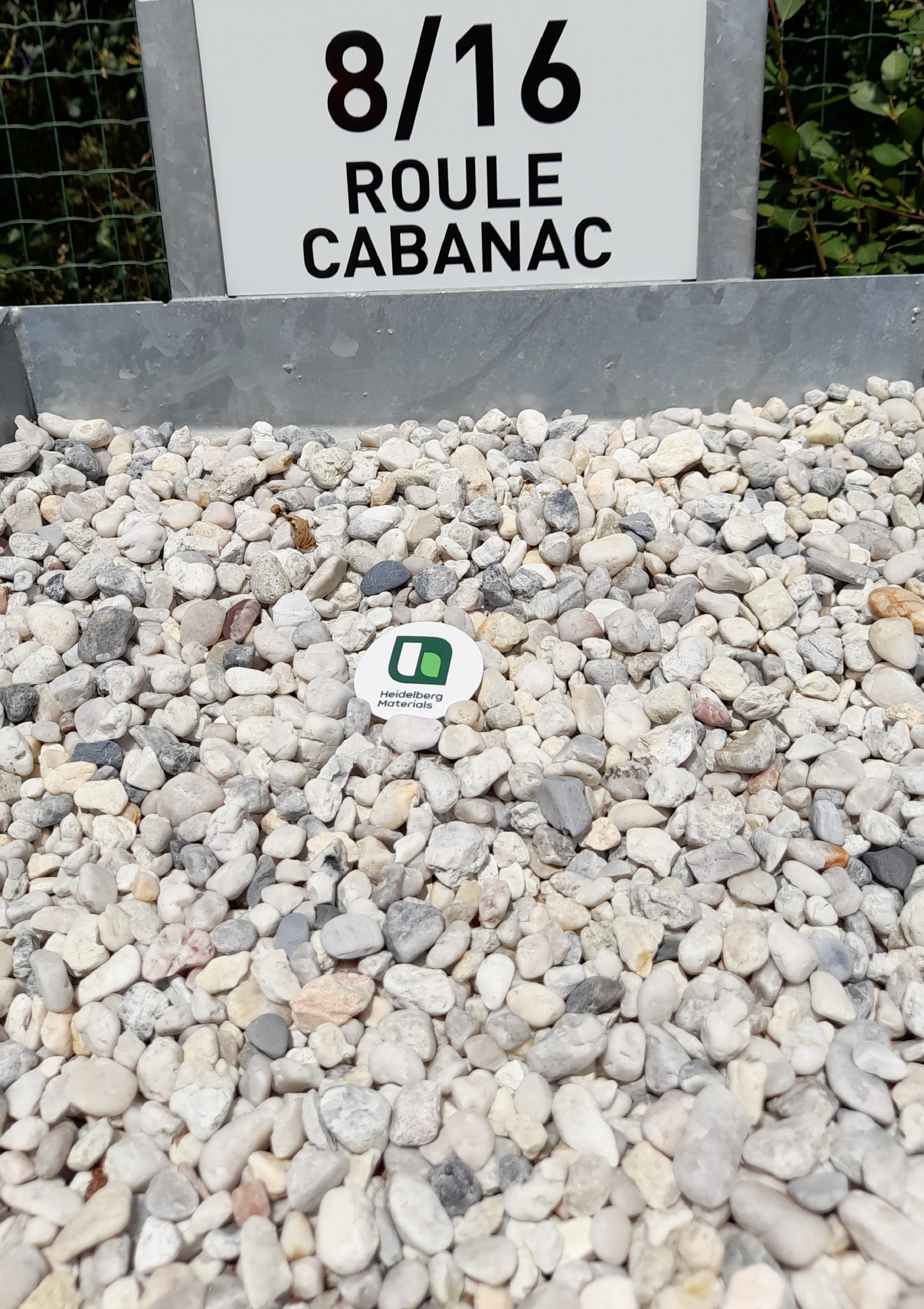 Plaque indiquant "8/16 Roule Cabanac" en haut d’un tas de graviers clairs, avec un marqueur rond vert et blanc posé au centre des graviers. Une clôture verte est visible en arrière-plan.