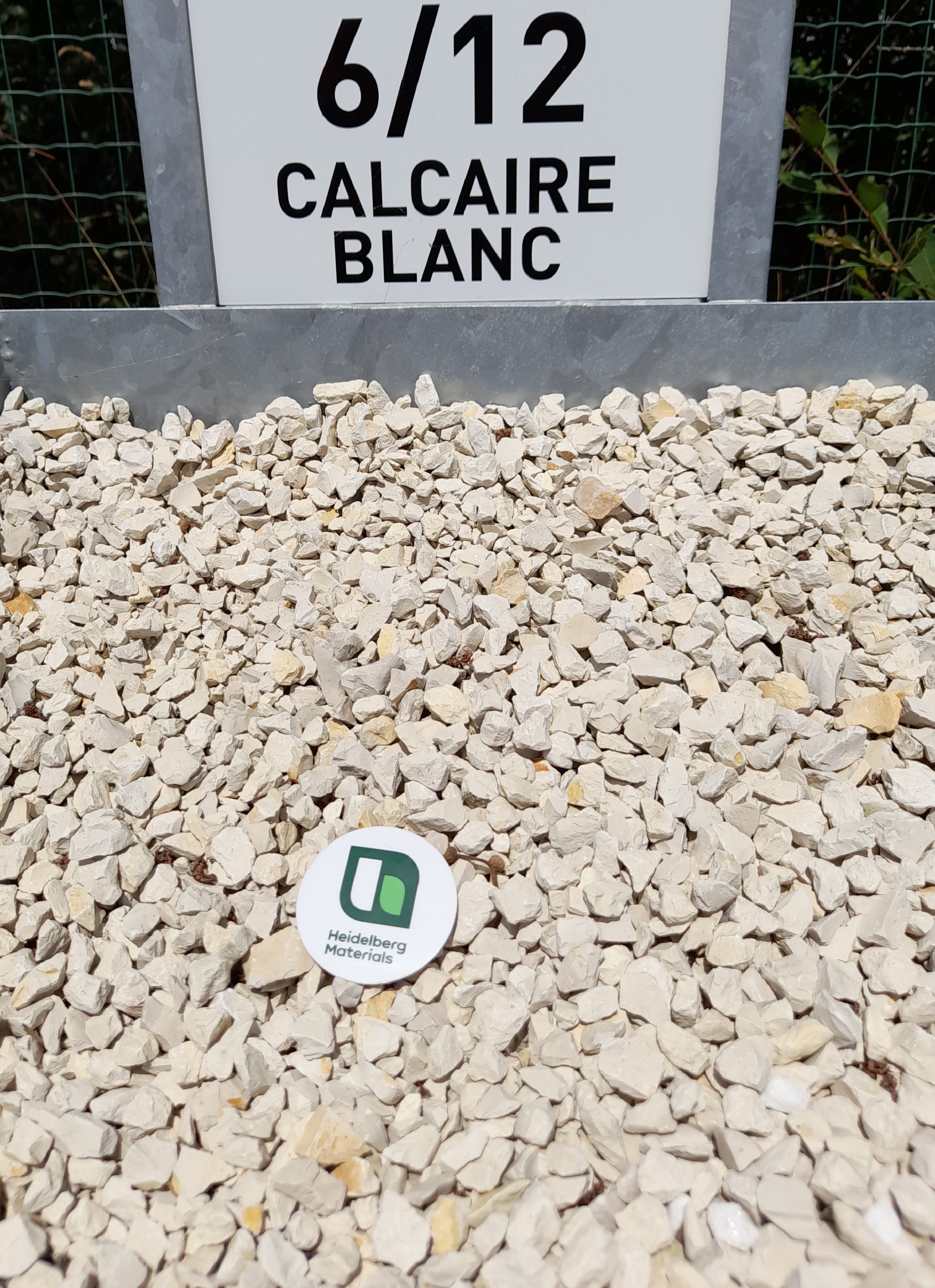 Gravier blanc de type calcaire 6/12 présenté en vrac, avec un panneau explicatif en arrière-plan affichant "6/12 CALCAIRE BLANC". Un repère rond au centre montre une échelle pour la taille des granulats.
