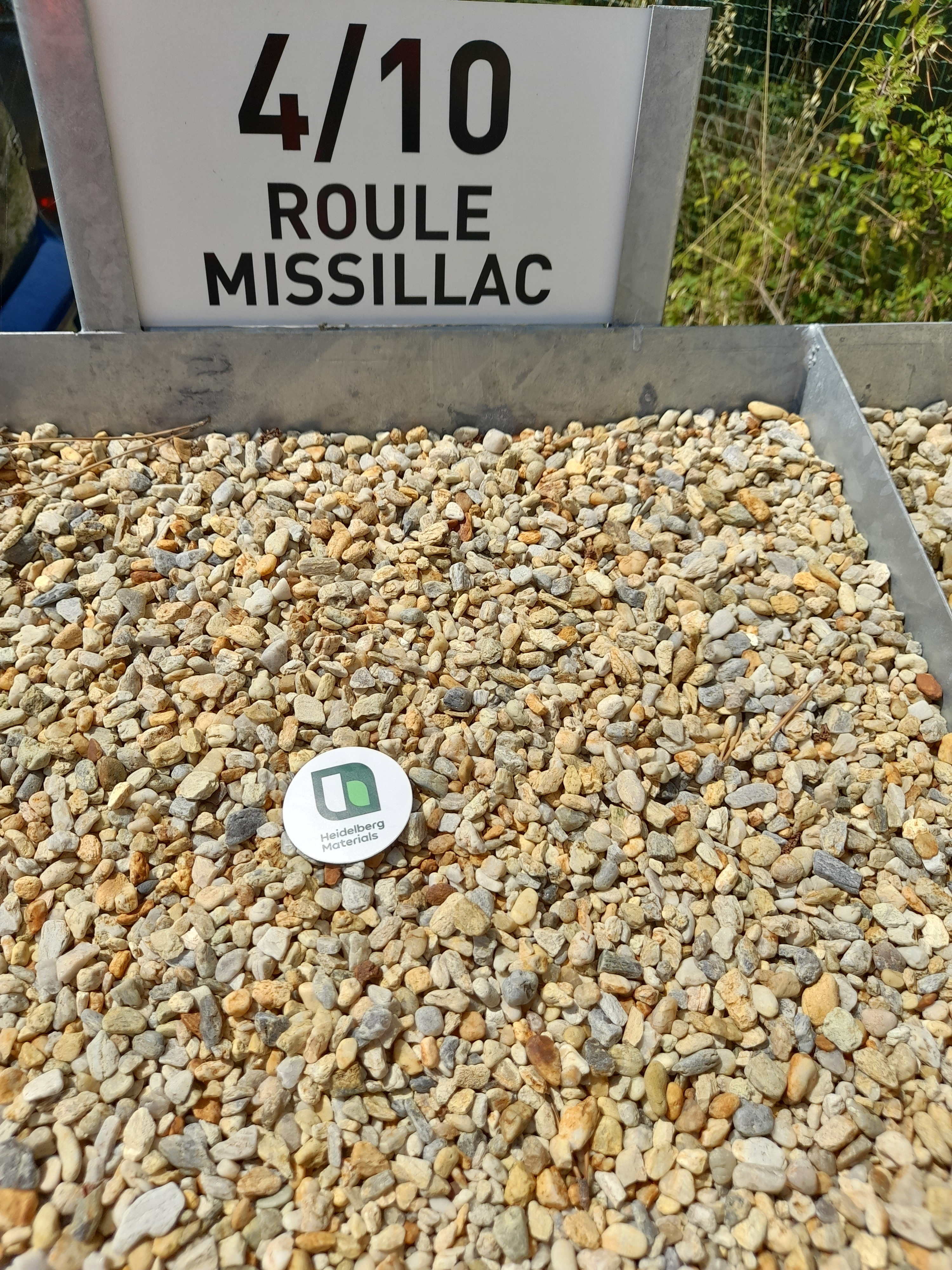 Gravier beige et brun clair disposé en bac, avec une étiquette circulaire contenant un code QR au centre. En arrière-plan, un panneau indiquant "4/10 ROULE MISSILLAC".