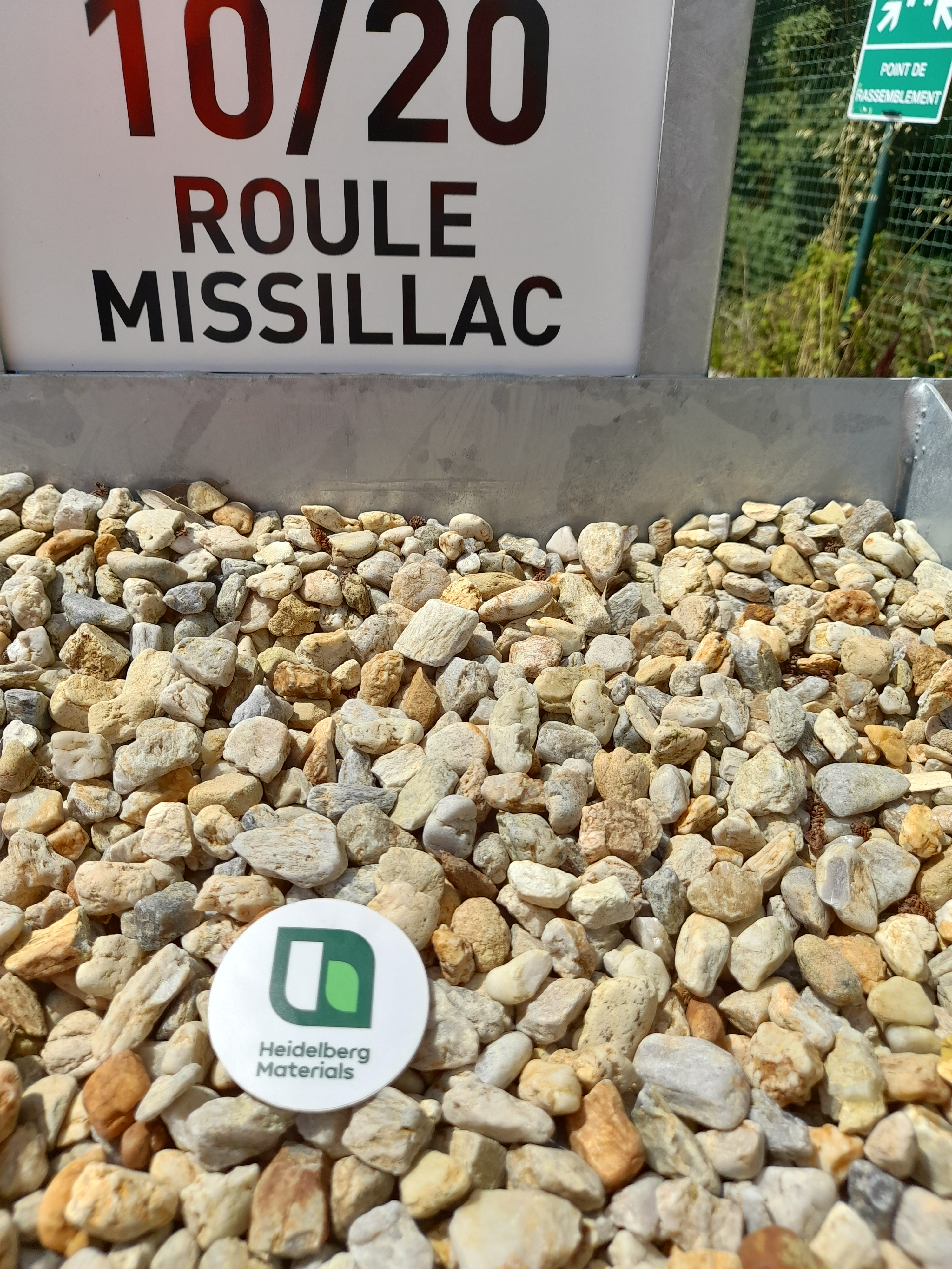Panneau affichant "10/20 Roule Missillac" en haut, posé au-dessus d’un lit de graviers avec un badge blanc portant le logo d’une mairie verte au centre. À droite, un panneau routier est partiellement visible sur un fond arboré.