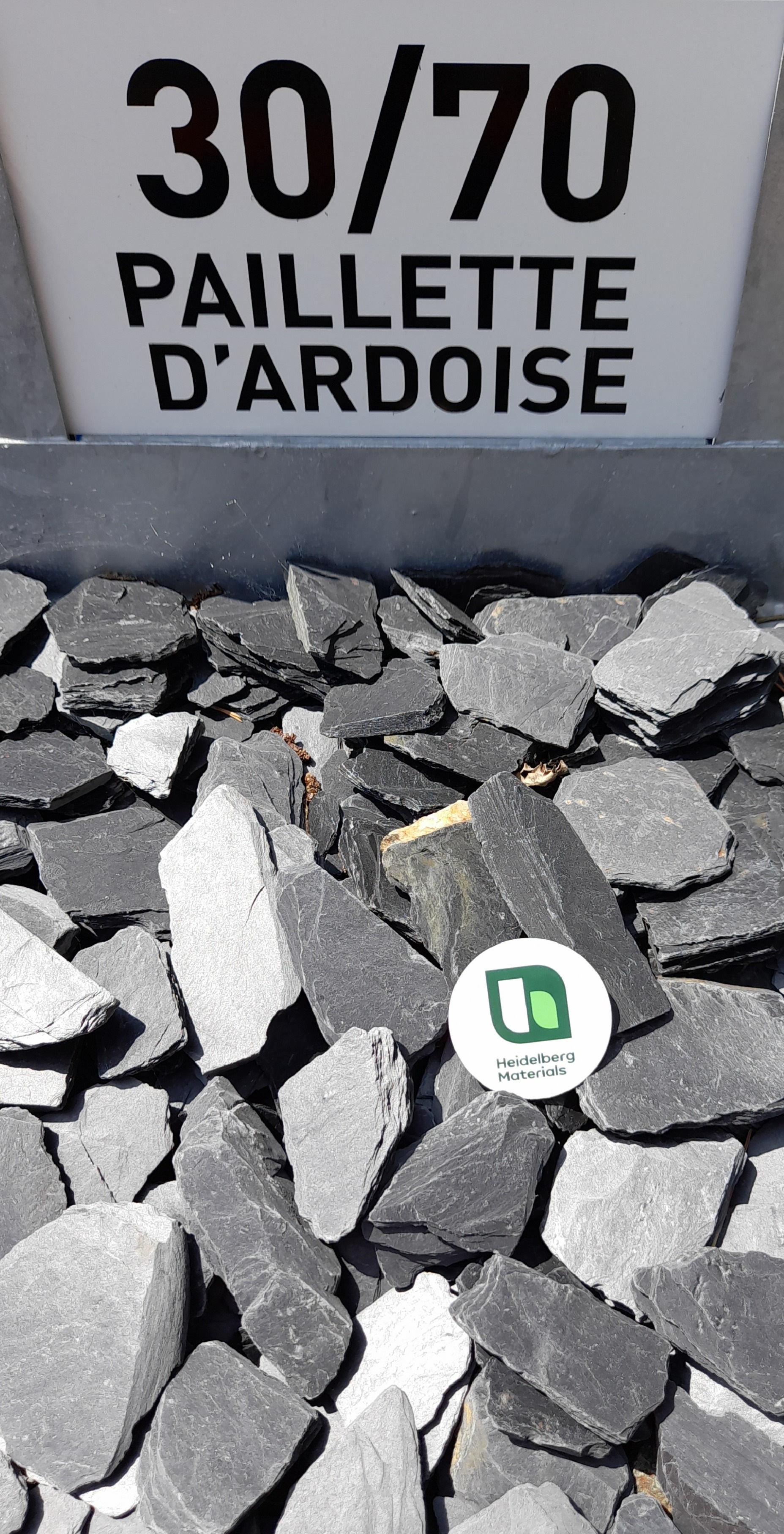 Plaquettes de schiste noir de taille 30-70 mm, disposées dans un bac d'exposition, avec un panneau descriptif "30/70 PAILLETTE D'ARDOISE" en arrière-plan. Un marqueur blanc rond avec un logo vert est placé au centre des pierres.