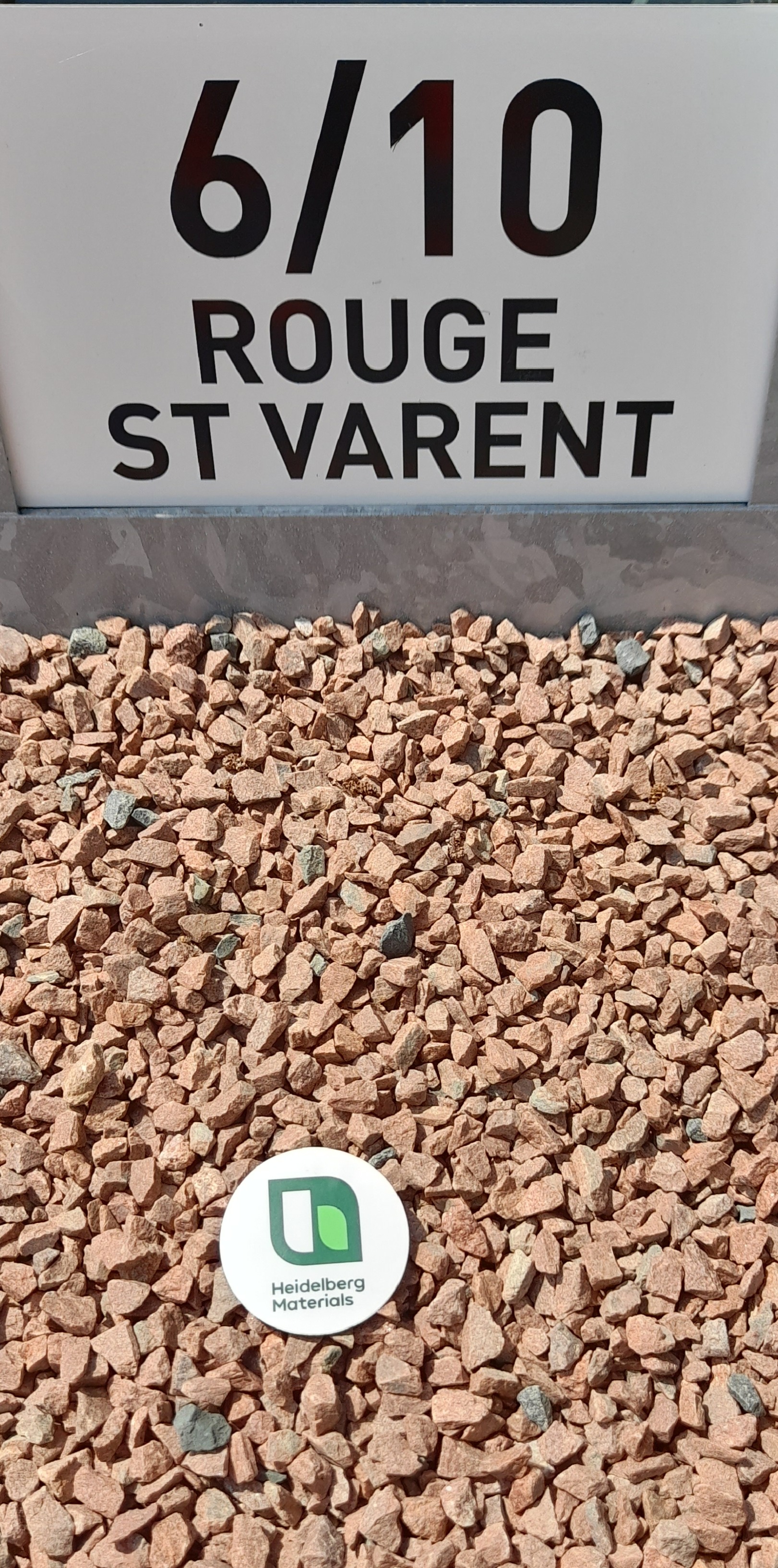 Granulats rouges de type "Rouge St Varent" de taille 6/10 mm, disposés sur le sol. En bas au centre, un disque blanc avec un logo vert et noir de la société productrice. À l’arrière-plan, un panneau blanc indiquant "6/10 Rouge St Varent".