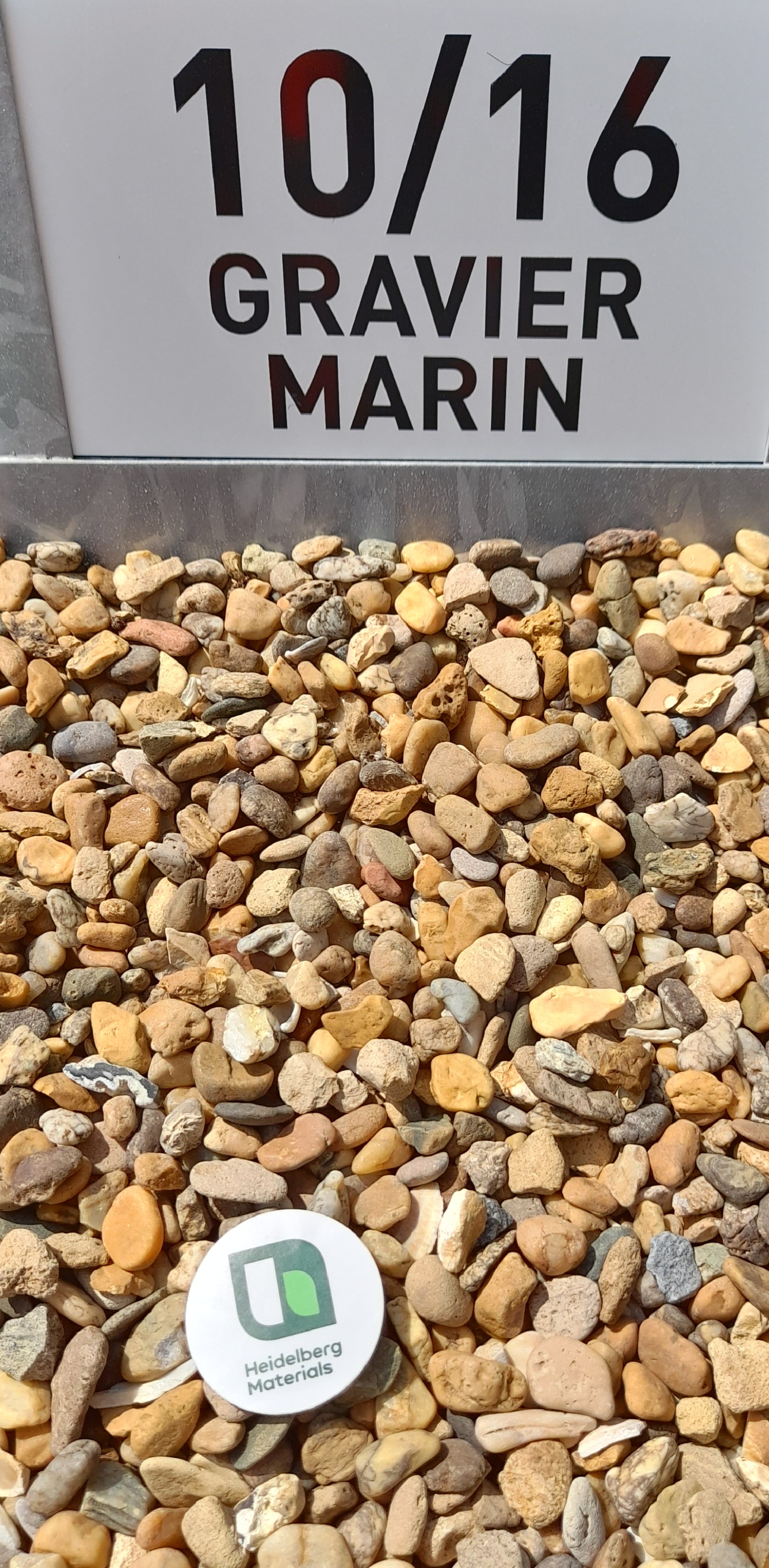 Gros plan de gravier marin de taille 10/16 mm avec un écriteau "10/16 GRAVIER MARIN" en arrière-plan. Un disque blanc avec un logo vert et gris est posé sur les graviers au premier plan.