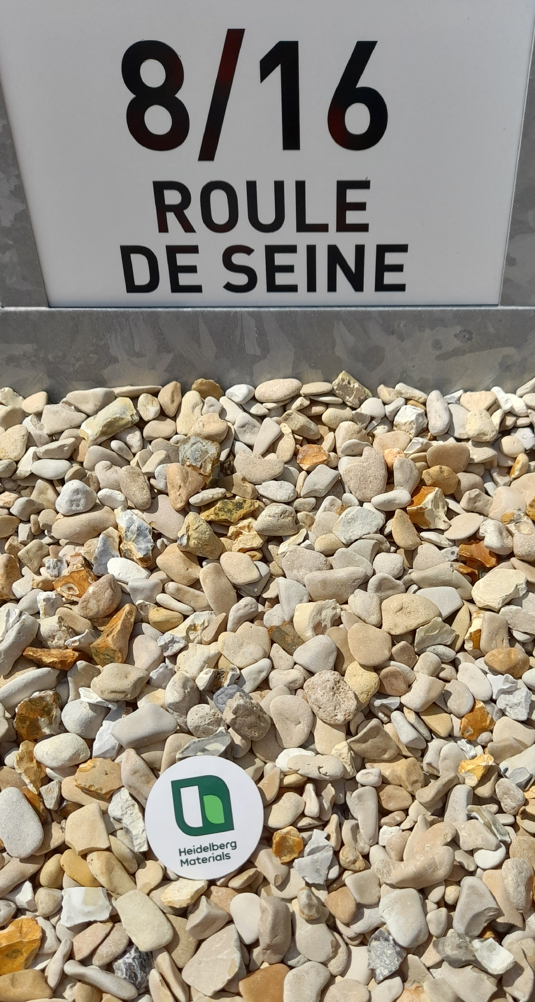 Gravillons beige clair et blanc marqués "8/16 Roule de Seine" dans une caisse de présentation, avec un badge rond portant le logo "u matériaux" posé parmi les pierres.