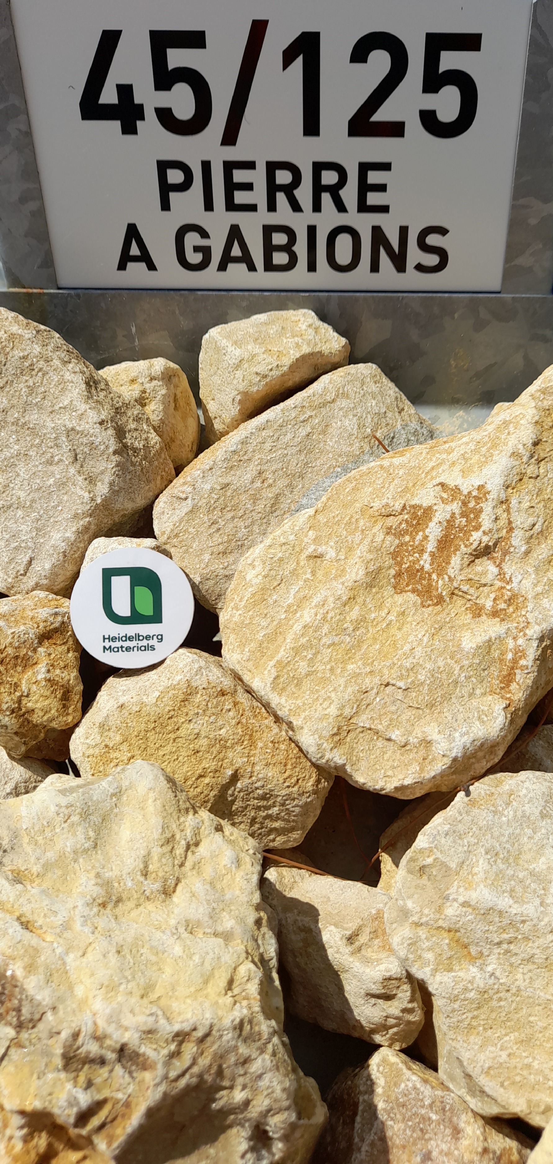 Pierres beige-jaune de taille 45/125 mm pour gabions, avec un panneau d'identification en haut indiquant "PIERRE À GABIONS" et une étiquette circulaire avec un logo vert au centre.