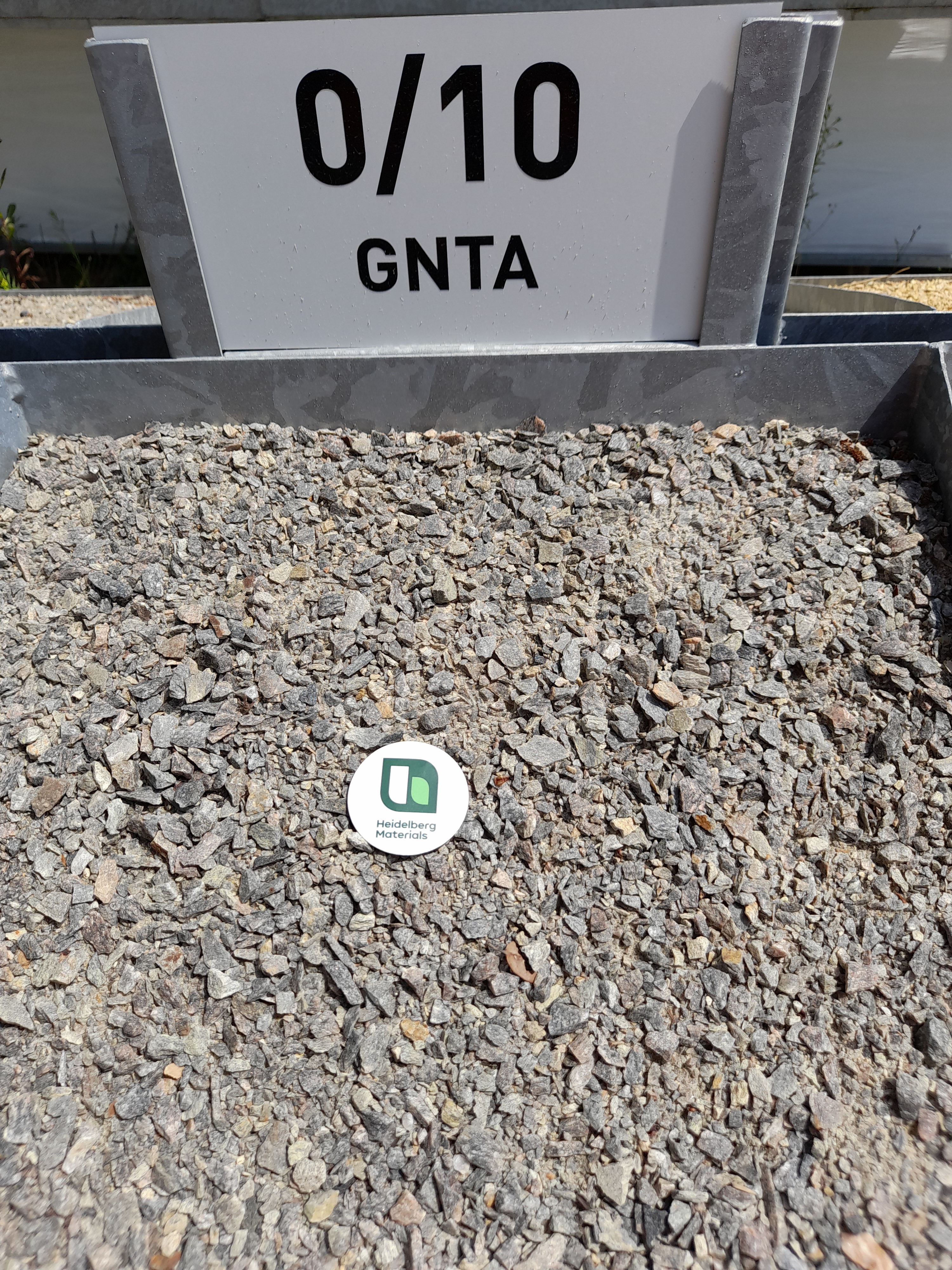 Tas de graviers fins, étiqueté "0/10 GNTA" en haut. Une pastille blanche avec un logo vert est posée sur les graviers au centre. L'arrière-plan montre un environnement extérieur flou.