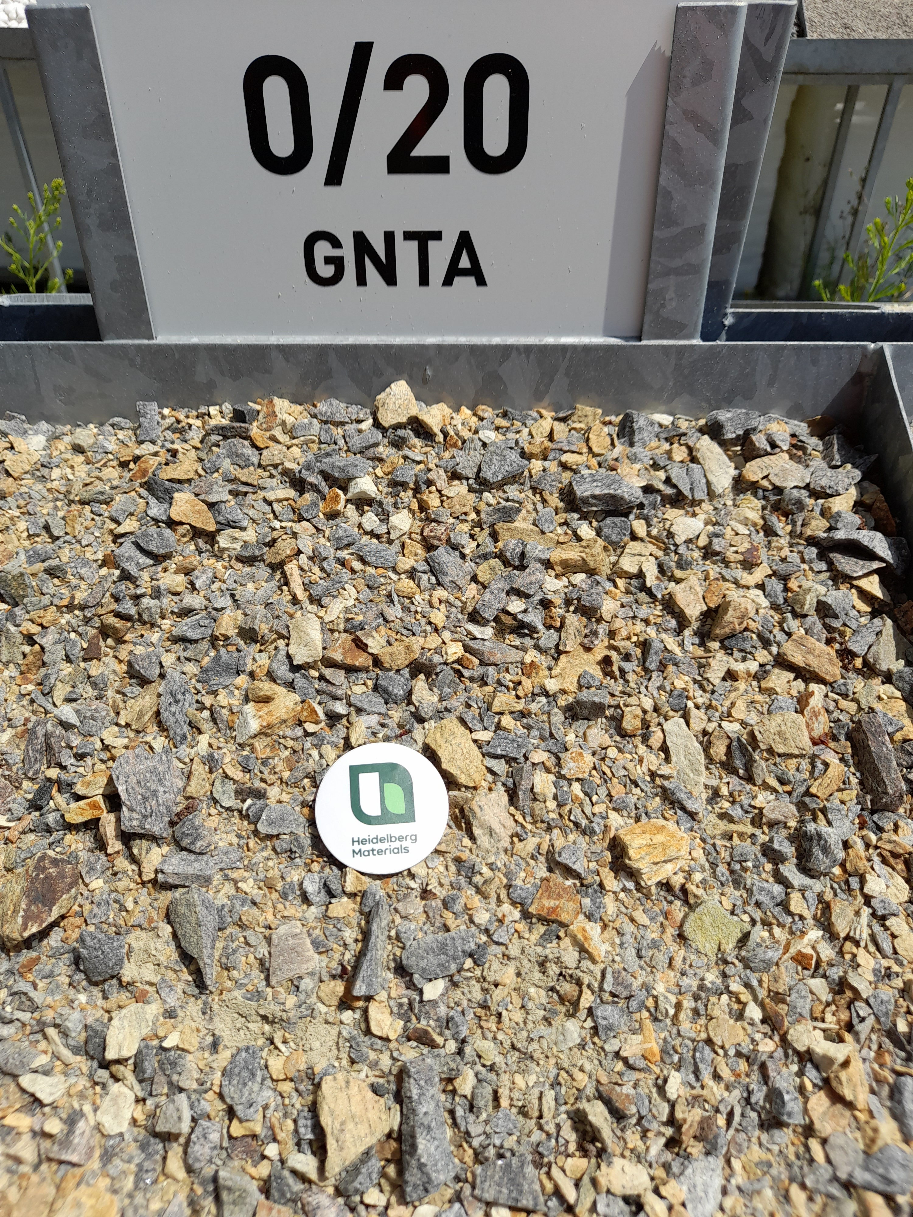 Gravier concassé de calibre 0/20 GNTA exposé dans un bac en métal, avec une étiquette ronde contenant un logo vert au centre. Panneau en arrière-plan indiquant "0/20 GNTA".
