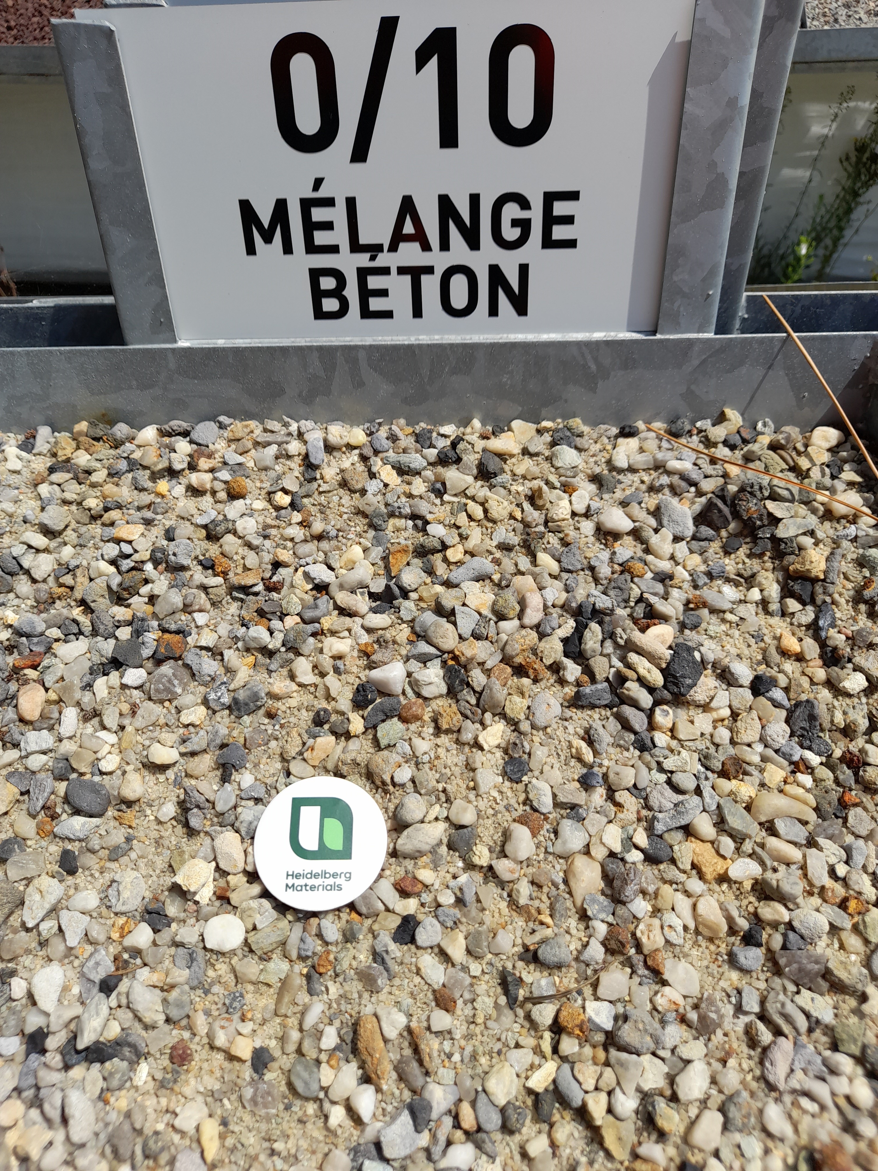Gravier mélangé de granulométrie 0/10 pour béton, présenté dans un bac à l'extérieur. Un logo circulaire vert et blanc est posé au centre des graviers.