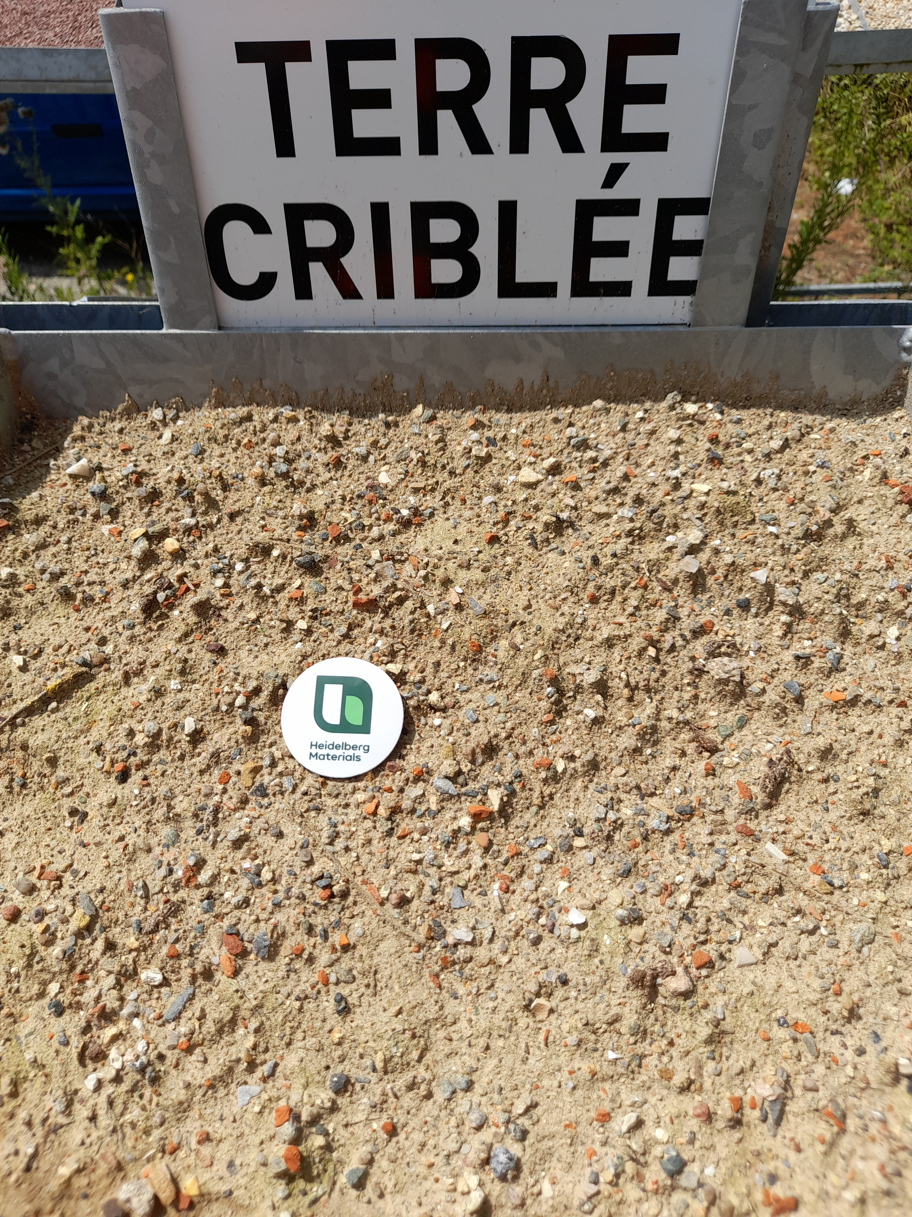 Tas de terre criblée avec une texture sableuse, contenant des petits cailloux. Un panneau blanc en arrière-plan indique "TERRE CRIBLÉE". Un disque blanc au centre affiche un logo vert.