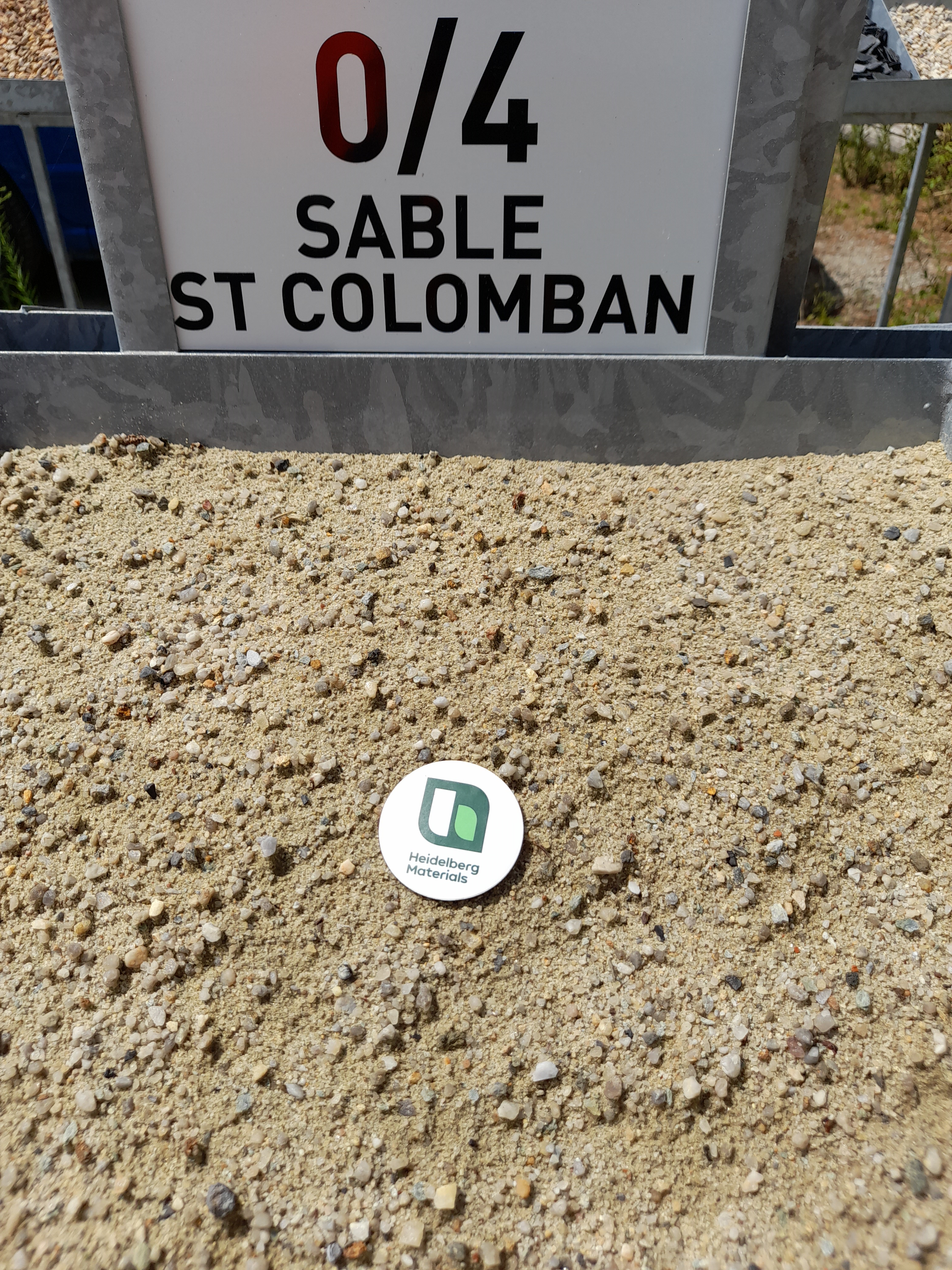 Pile de sable fin et granuleux étiqueté "0/4 Sable St Colomban" à l’arrière-plan, avec un badge circulaire blanc montrant un logo vert posé sur le sable au centre-bas de l’image.