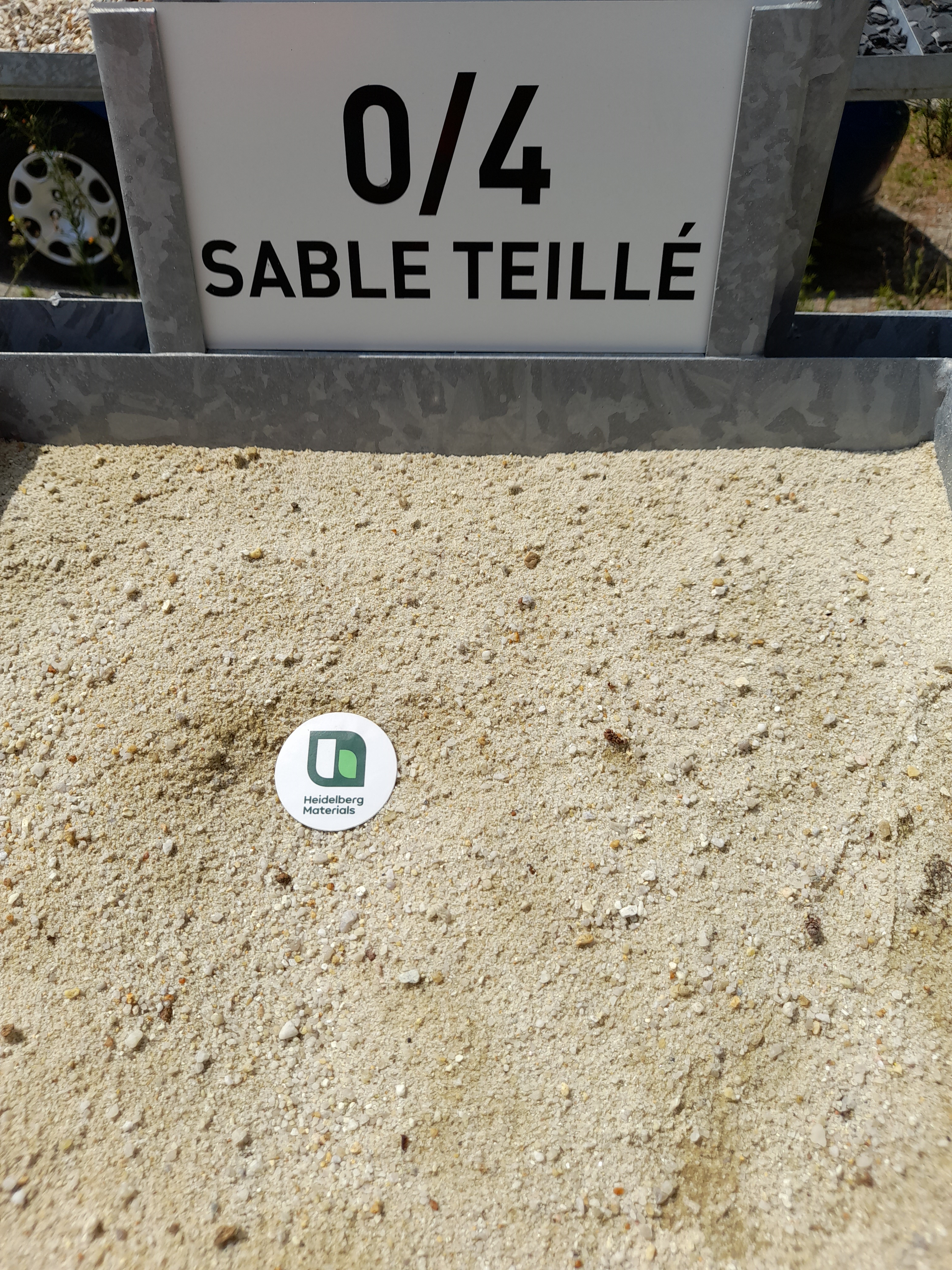 Tas de sable fin de granulométrie 0/4 (sable teillé), clairement étiqueté sur un panneau au-dessus. Une étiquette ronde avec un logo vert est posée sur le sable, indiquant un éventuel marquage ou certification.