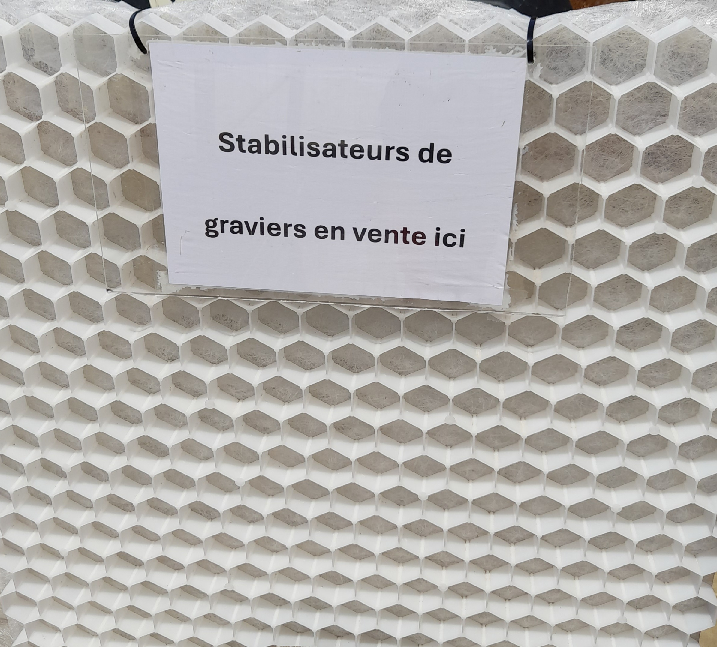 Panneau affichant "Stabilisateurs de graviers en vente ici" fixé sur une structure alvéolée blanche en plastique, utilisée pour stabiliser les graviers.