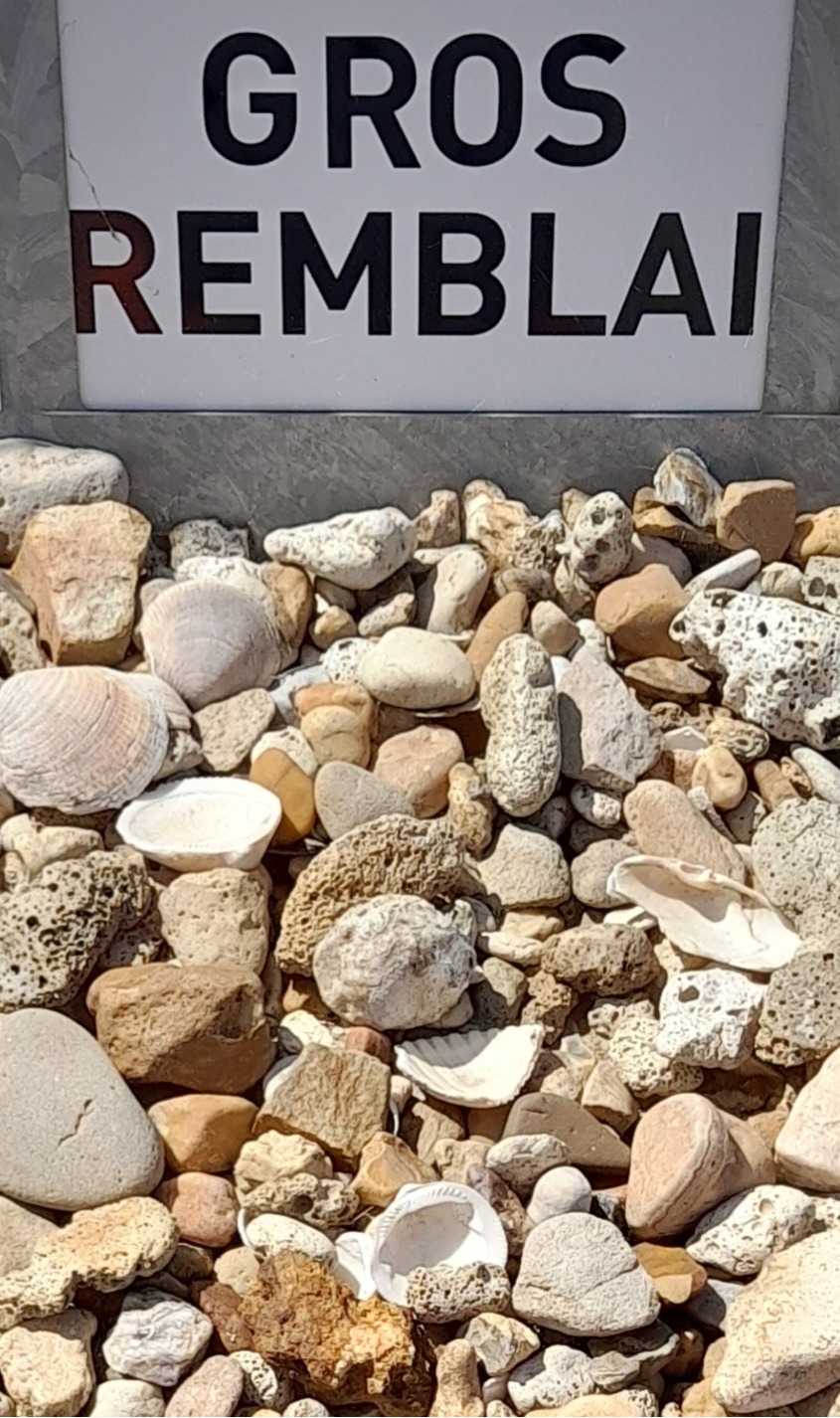 Pile de gros cailloux et fragments de roche beige et blanche au sol, accompagnée d'un panneau blanc en arrière-plan inscrit "GROS REMBLAI" en lettres noires et rouges.