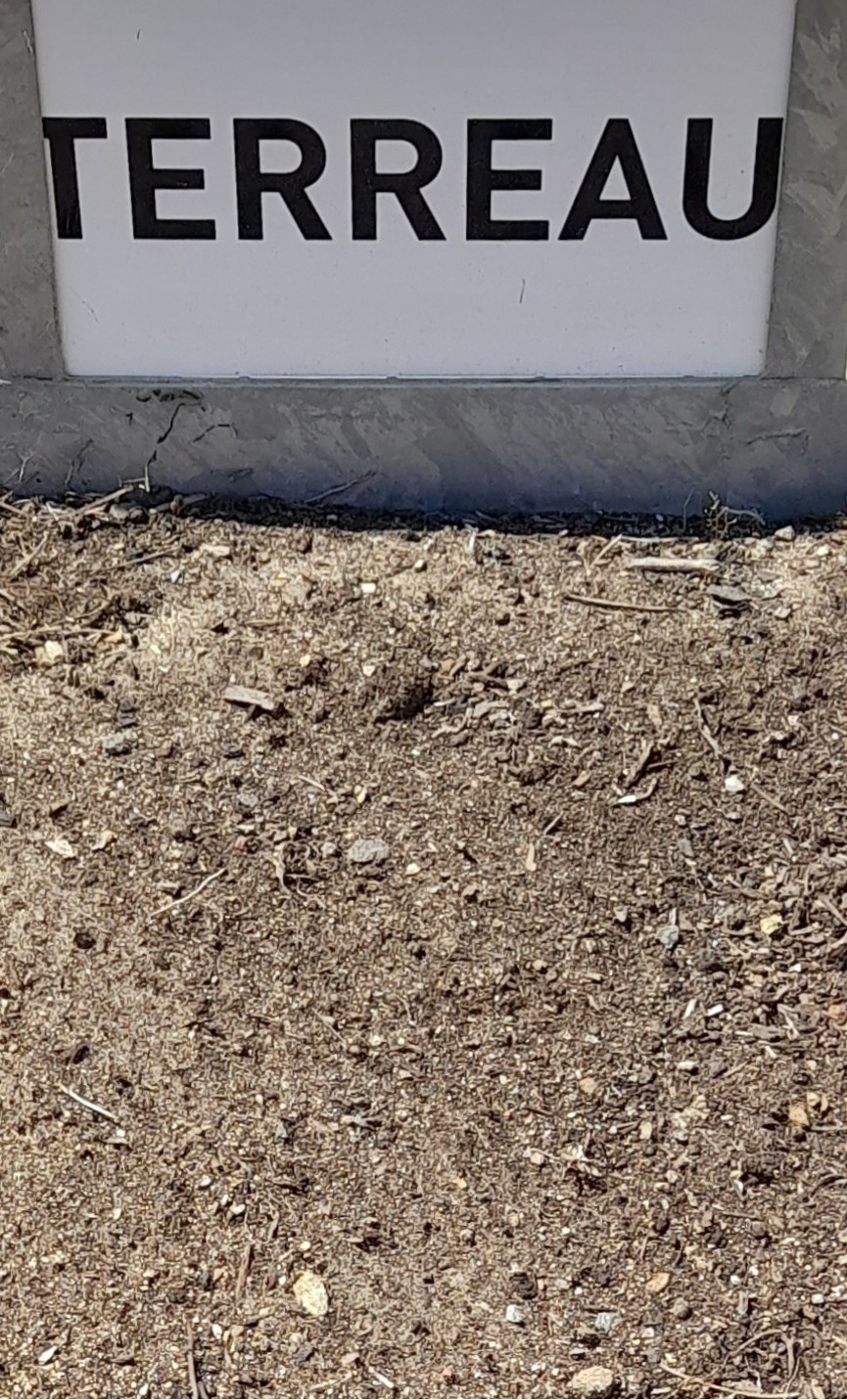 Tas de terreau brun devant un panneau métallique avec l'inscription "TERREAU" en lettres noires, indiquant un emplacement dédié au terreau.