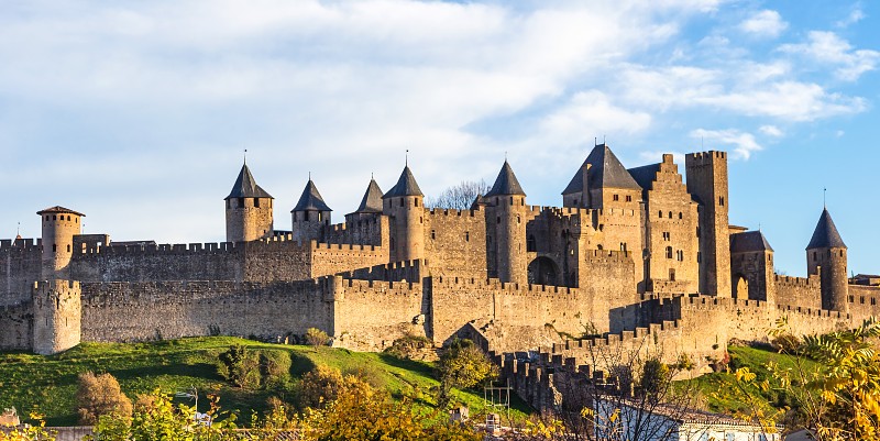 Medieval fortress with multiple towers and stone walls on a grassy hill under a blue sky.
